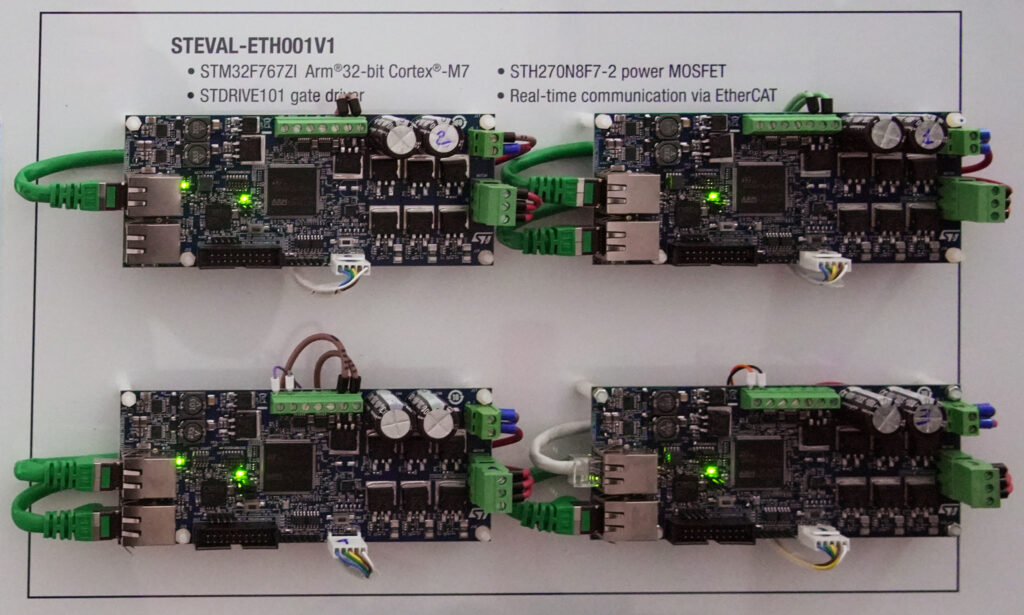 Vista dall'alto di quattro schede elettroniche STMicroelectronics STEVAL-ETH001V1, montate su una superficie bianca. Le schede includono componenti come il microcontrollore STM32F767ZI, driver gate STDRIVE101 e MOSFET di potenza STH270N8F7-2. Sono collegate tramite cavi Ethernet verdi e altri cavi di connessione, con alcuni LED verdi accesi che indicano il funzionamento.