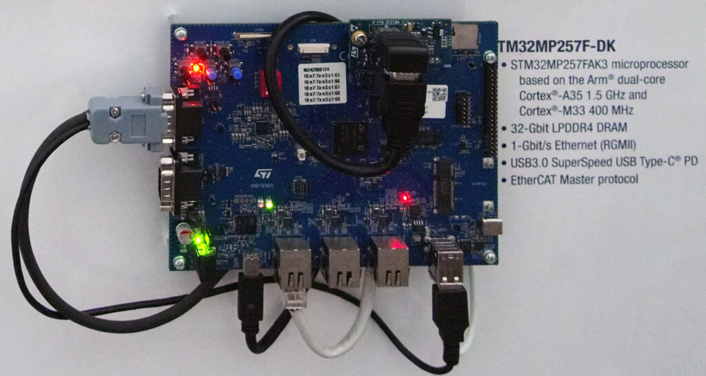 Vista dall'alto di una scheda elettronica STMicroelectronics STM32MP257F-DK, montata su una superficie bianca. La scheda include un microprocessore STM32MP257FAK3 basato su Arm® dual-core Cortex®-A35 a 1,5 GHz e Cortex®-M33 a 400 MHz, 32-Gbit LPDDR4 DRAM, Ethernet 1-Gbit/s (RGMII), USB 3.0 SuperSpeed USB Type-C® PD e protocollo EtherCAT Master. Sono collegati vari cavi, inclusi cavi Ethernet e USB, con alcuni LED verdi e rossi accesi che indicano il funzionamento.