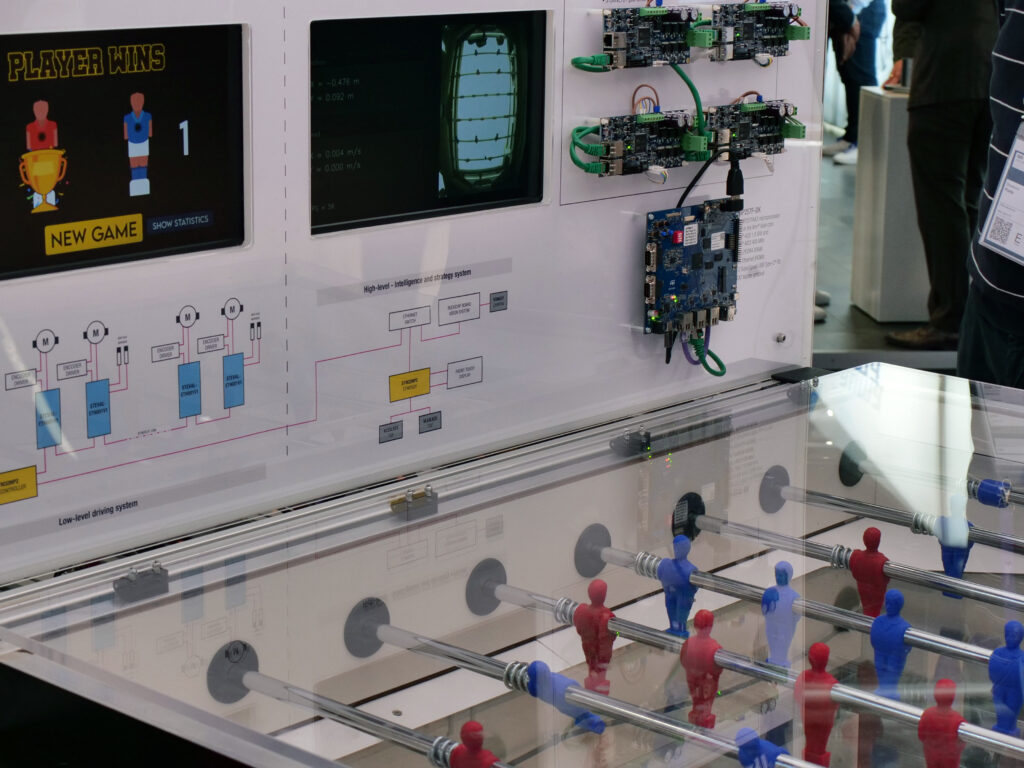Immagine di un calcetto robotizzato in esposizione. La parte superiore mostra due schermi: uno con il messaggio 'PLAYER WINS' e un altro con dati di gioco. Sulla destra, sono visibili diverse schede elettroniche STMicroelectronics montate su una superficie bianca. La parte inferiore dell'immagine mostra il campo da gioco del calcetto con omini rossi e blu. Sullo sfondo, si intravedono alcune persone.